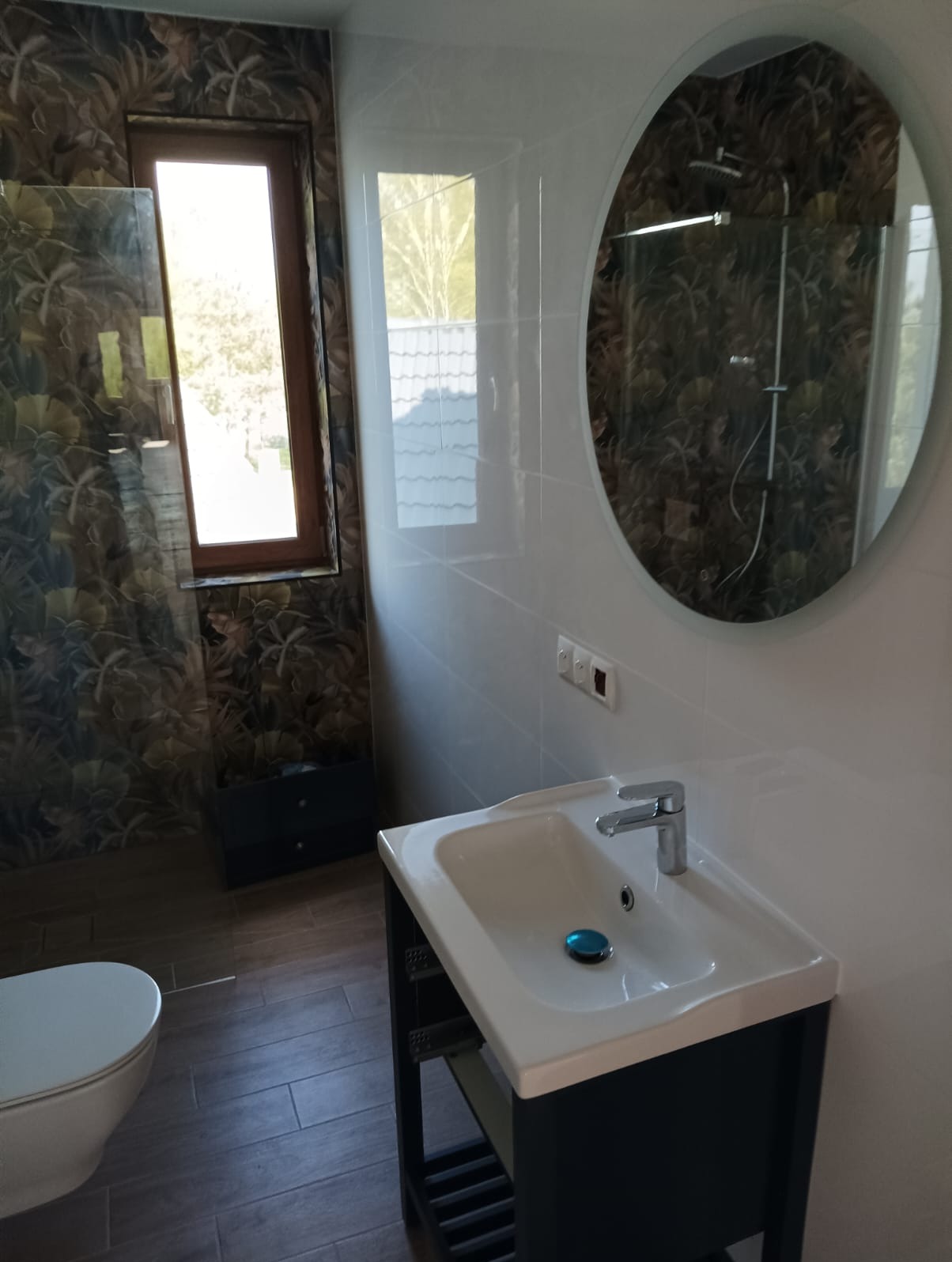 Wykończona łazienka z umywalką na szafce, okrągłym lustrem i deszczownicą w kabinie. Wnętrze zdobią płytki z motywem liści i drewnopodobna podłoga.