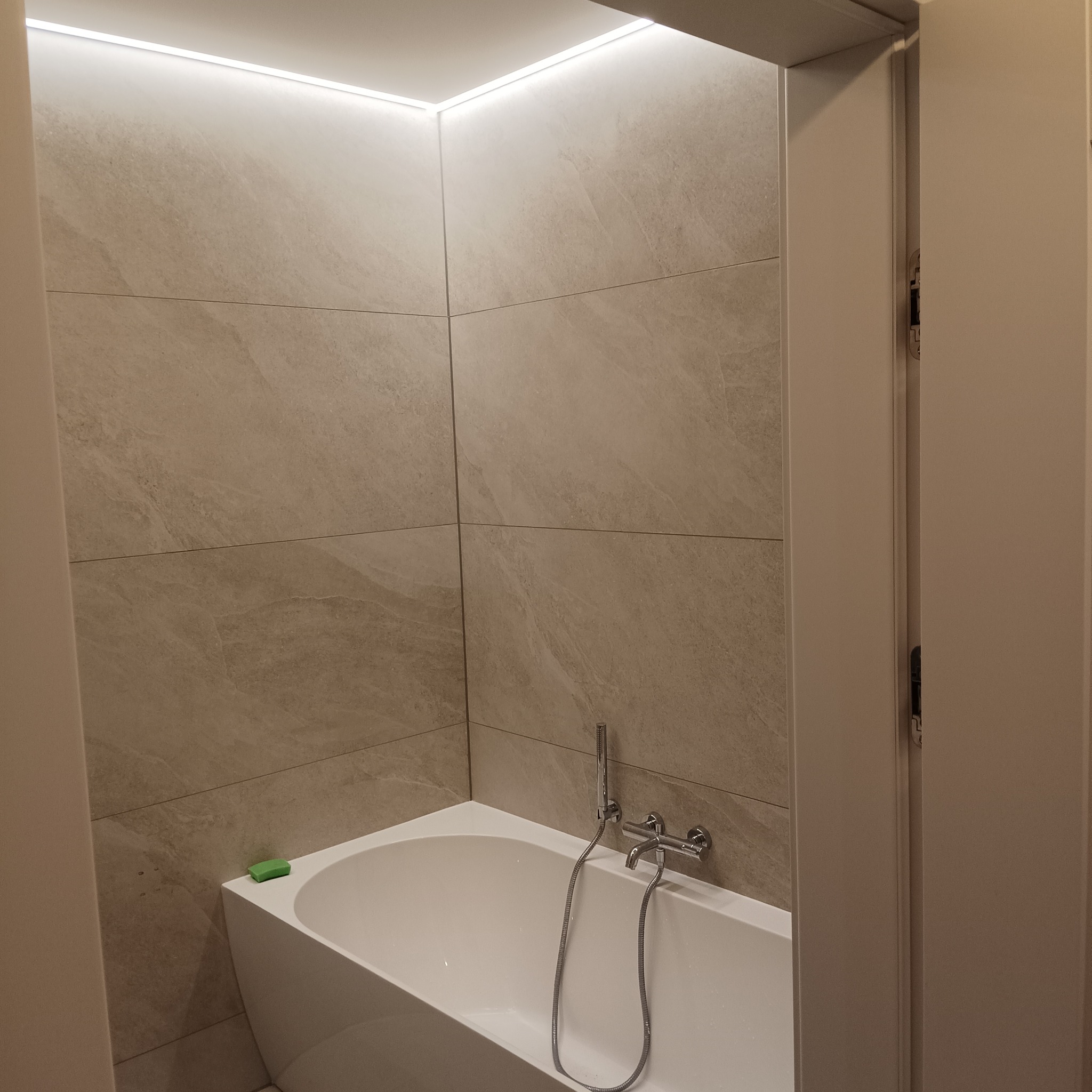 Nowoczesna łazienka z wanną, wykończona płytkami w odcieniach beżu. Oświetlenie LED podkreśla minimalistyczny design. Na wannie leży zielone mydło.