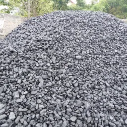 Góra ciemnego węgla kamiennego, drobnoziarnistego, ułożona przy betonowym murku, z zielenią w tle.