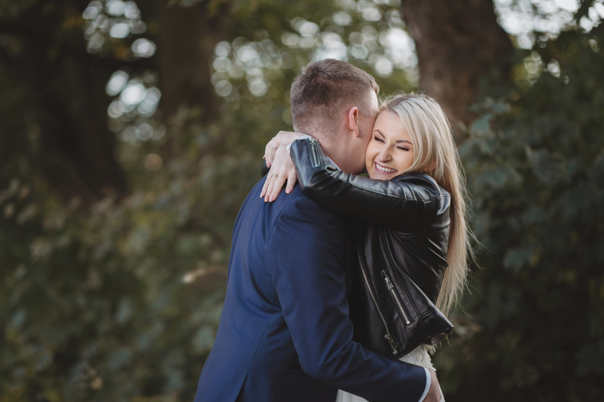 Uśmiechnięta blondynka w czarnej skórzanej kurtce obejmuje mężczyznę w granatowym garniturze na tle zieleni.