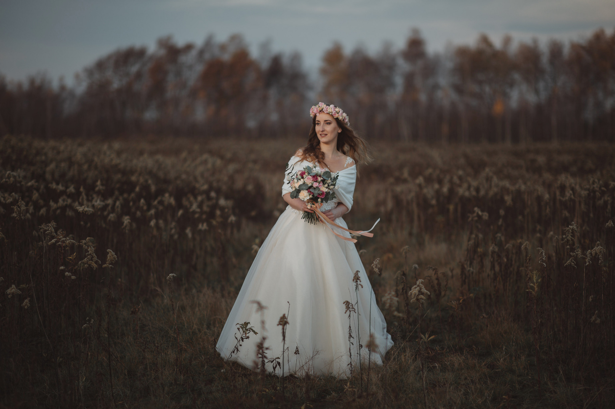 Panna młoda w białej sukni i wianku z różowych kwiatów, trzymająca bukiet, pozuje na tle łąki z wysoką trawą i drzewami w oddali.