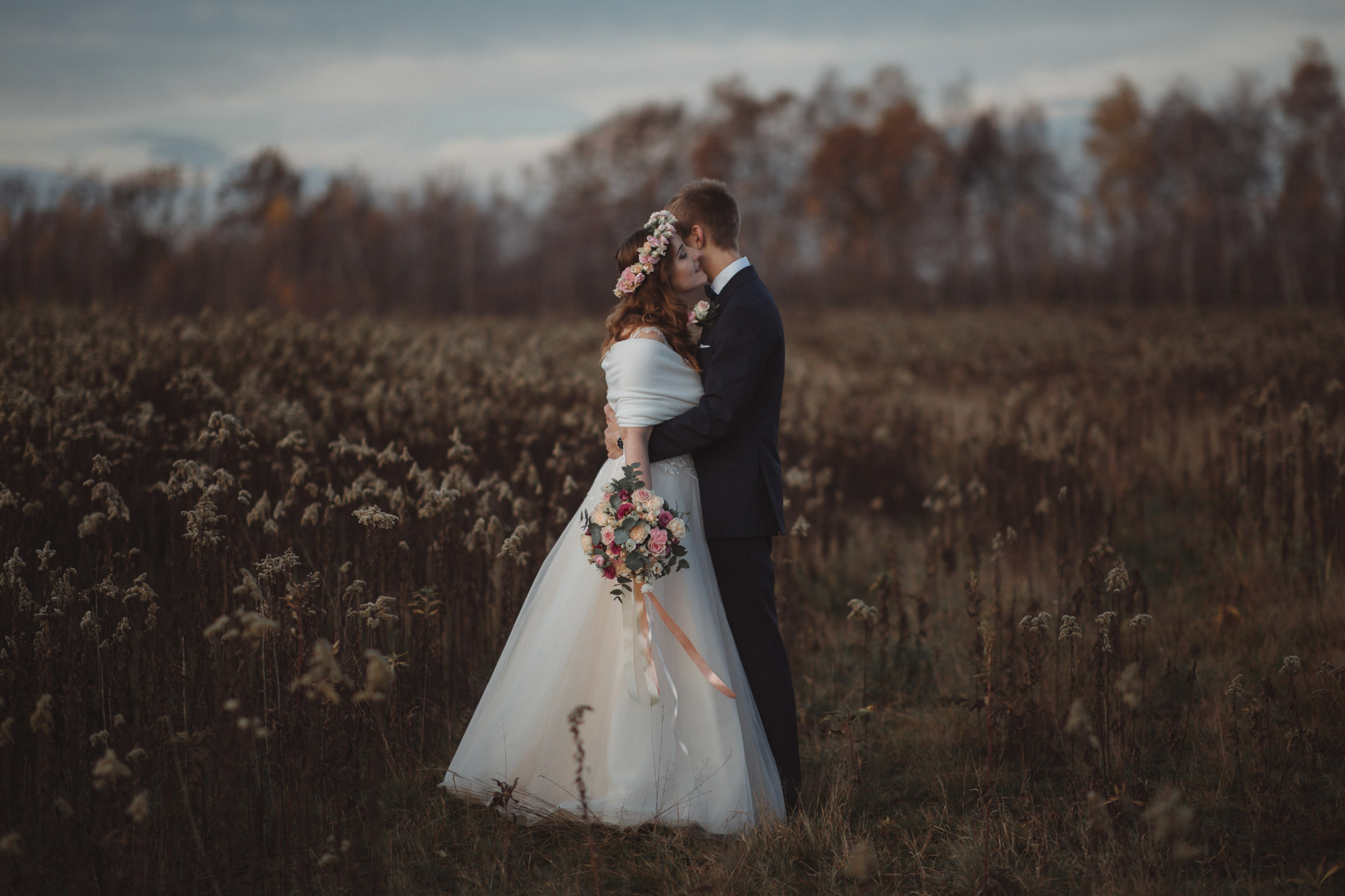 Para młoda w objęciach na tle jesiennego pola i drzew, panna młoda w sukni z kwiatowym wiankiem i bukietem, pan młody w garniturze.