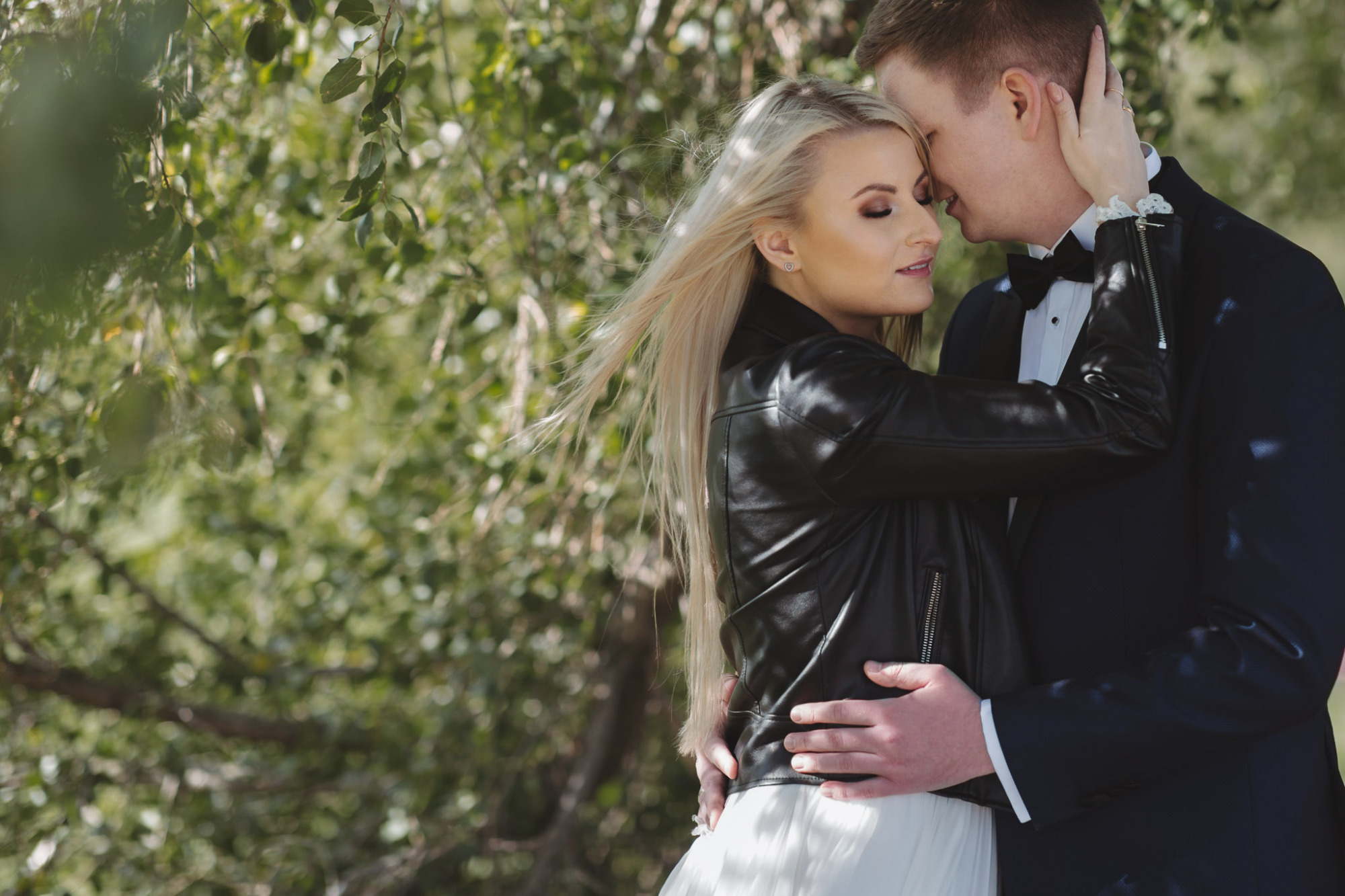Para młoda w objęciach na tle drzew, panna młoda w skórzanej kurtce i z wiatrem we włosach.