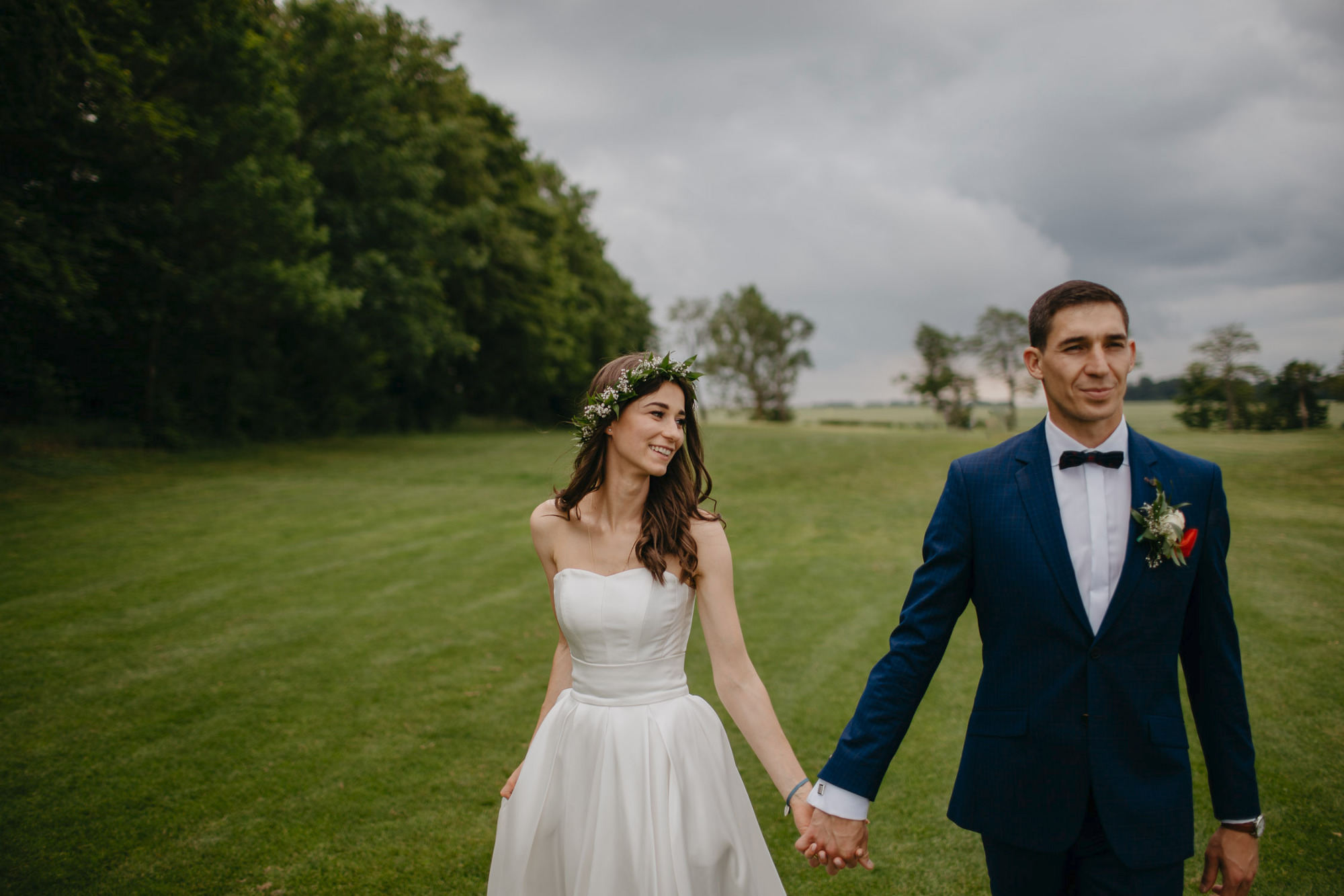 Para młoda trzymająca się za ręce spaceruje po zielonym polu, panna młoda w białej sukni i wianku, pan młody w granatowym garniturze z muchą.