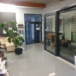 Ekspozycja w salonie: przesuwne drzwi tarasowe z czarną ramą, widoczne przez szybę rolety okienne, obok doniczka z rośliną, w tle sofa i próbki profili.