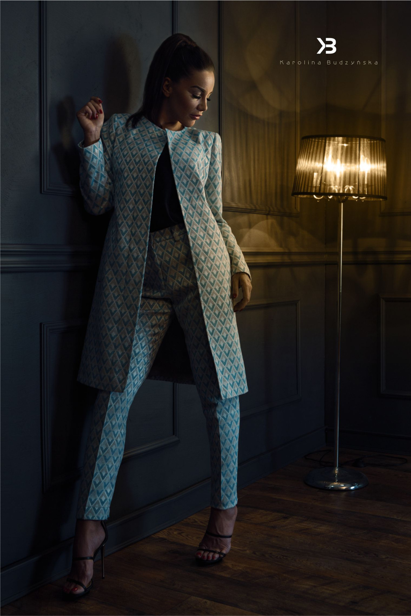 Kobieta w dopasowanym, wzorzystym komplecie (płaszcz i spodnie) w odcieniach błękitu i beżu, oparta o ciemną ścianę, obok lampa podłogowa, prezentacja kolekcji.