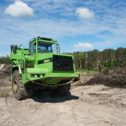 Zielony wozidło przegubowe Terex na placu budowy z koparką w tle, pod błękitnym niebem z lekkimi chmurami.