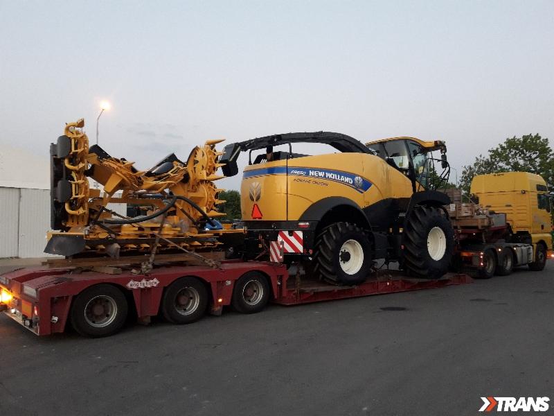 Ciężarówka z naczepą przewożąca żółty kombajn New Holland FR950 i przystawkę do kukurydzy na niskopodwoziowej platformie, widoczne zabezpieczenia ładunku.
