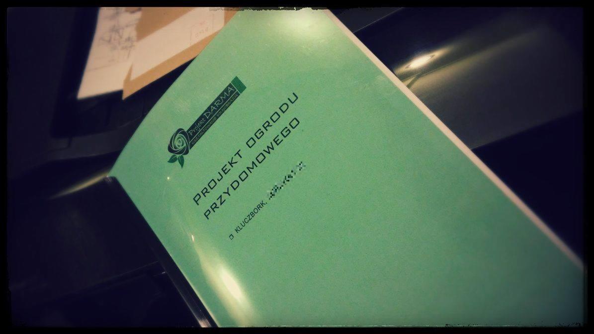 Zielona teczka z napisem 'Projekt ogrodu przydomowego' oraz logo firmy Projekt Darma, widoczna na tle biurka i innych dokumentów.