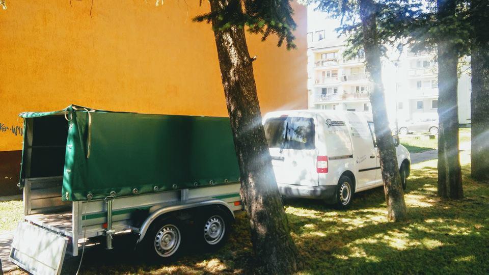 Przyczepa z zieloną plandeką i biały van zaparkowane obok budynku, widoczne drzewa i trawa w słoneczny dzień.