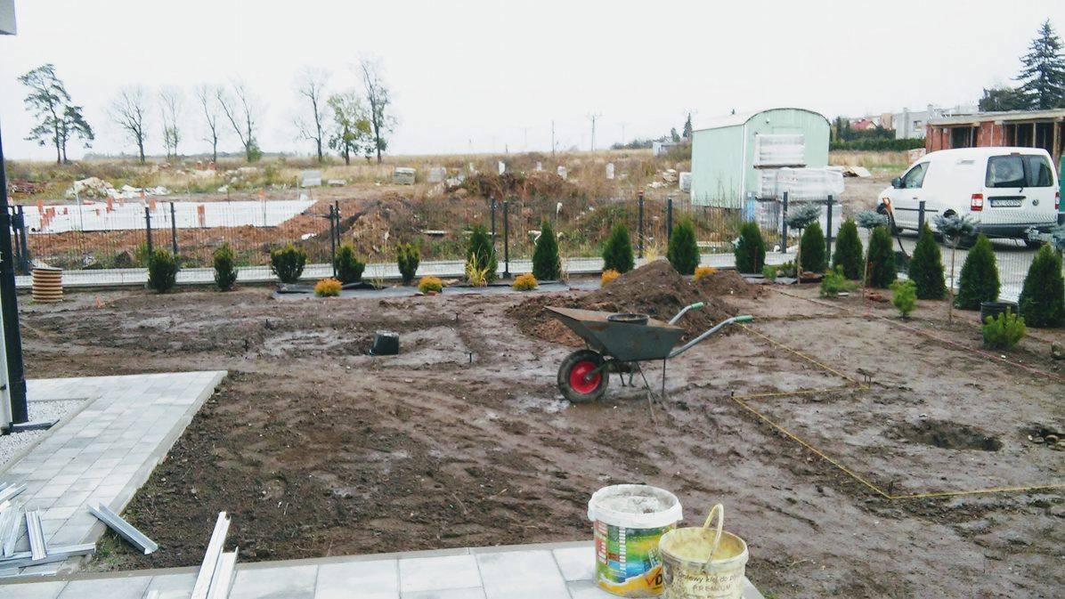 Wykopana działka z przygotowanymi nasadzeniami iglaków i krzewów ozdobnych, taczka z ziemią, obrys fundamentów i widoczny w tle samochód dostawczy.