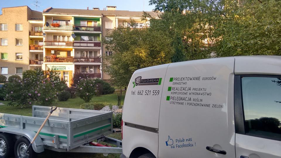 Samochód firmy ogrodniczej z Kluczborka z przyczepą i narzędziami na tle bloku mieszkalnego, widoczne logo i numer telefonu firmy.