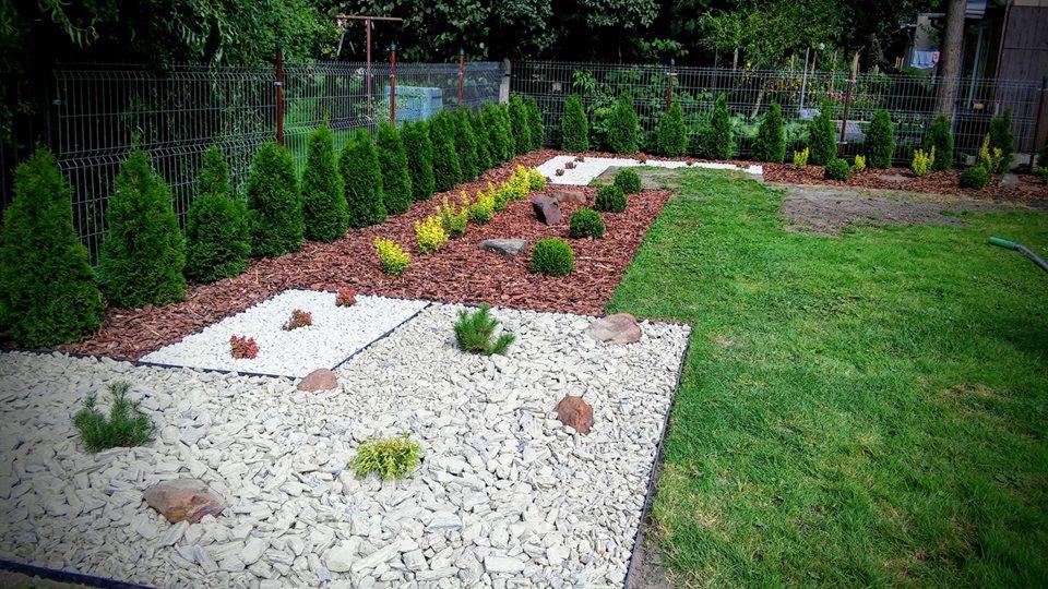 Uporządkowany ogród z rabatami wysypanymi korą i białym kamieniem, obrzeża z tuj i trawnika, widoczne pojedyncze kamienie dekoracyjne.