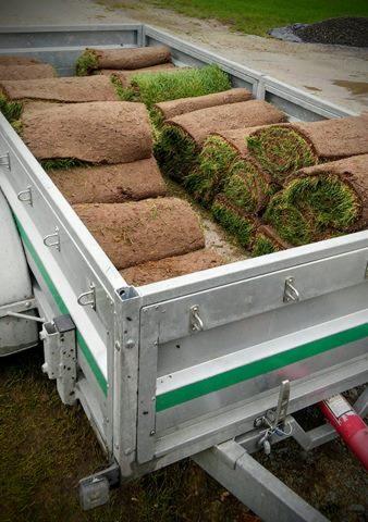 Przyczepa wypełniona rolkami świeżej, zwiniętej darni trawnikowej, gotowa do rozłożenia, widoczne fragmenty zielonej trawy i brązowej ziemi.