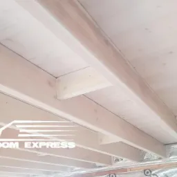 Wnętrze domu w konstrukcji szkieletowej, widoczne drewniane belki stropowe i ocieplenie ścian folią aluminiową, logo firmy w lewym dolnym rogu.