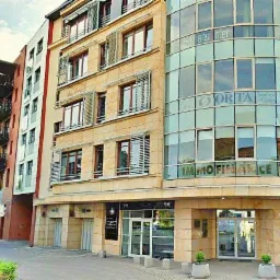 Widok z zewnątrz na nowoczesny budynek biurowy z przeszkloną fasadą, na której widnieją nazwy firm, w tym Immo Finance i ORITA, z widocznymi tablicami informacyjnymi i zaparkowanymi samochodami.