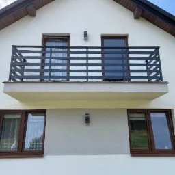 Elewacja budynku z balkonem zabezpieczonym nowoczesną, poziomą, metalową konstrukcją, widoczne okna z brązowymi ramami i oświetlenie zewnętrzne.