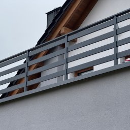 Adamit - Szara, pozioma balustrada balkonowa z drewnianymi elementami, widok z dołu na tle elewacji i fragmentu dachu domu.