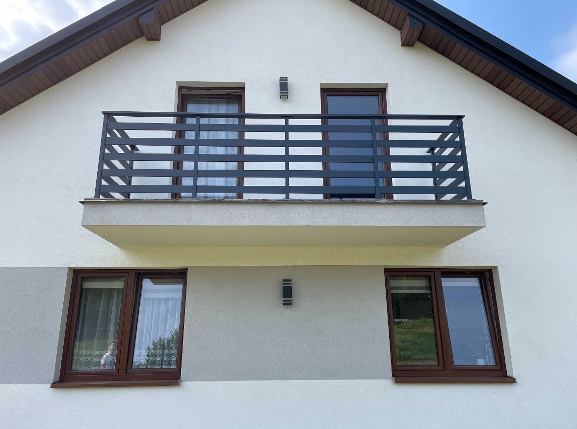 Elewacja budynku z balkonem zabezpieczonym nowoczesną, poziomą, metalową konstrukcją, widoczne okna z brązowymi ramami i oświetlenie zewnętrzne.