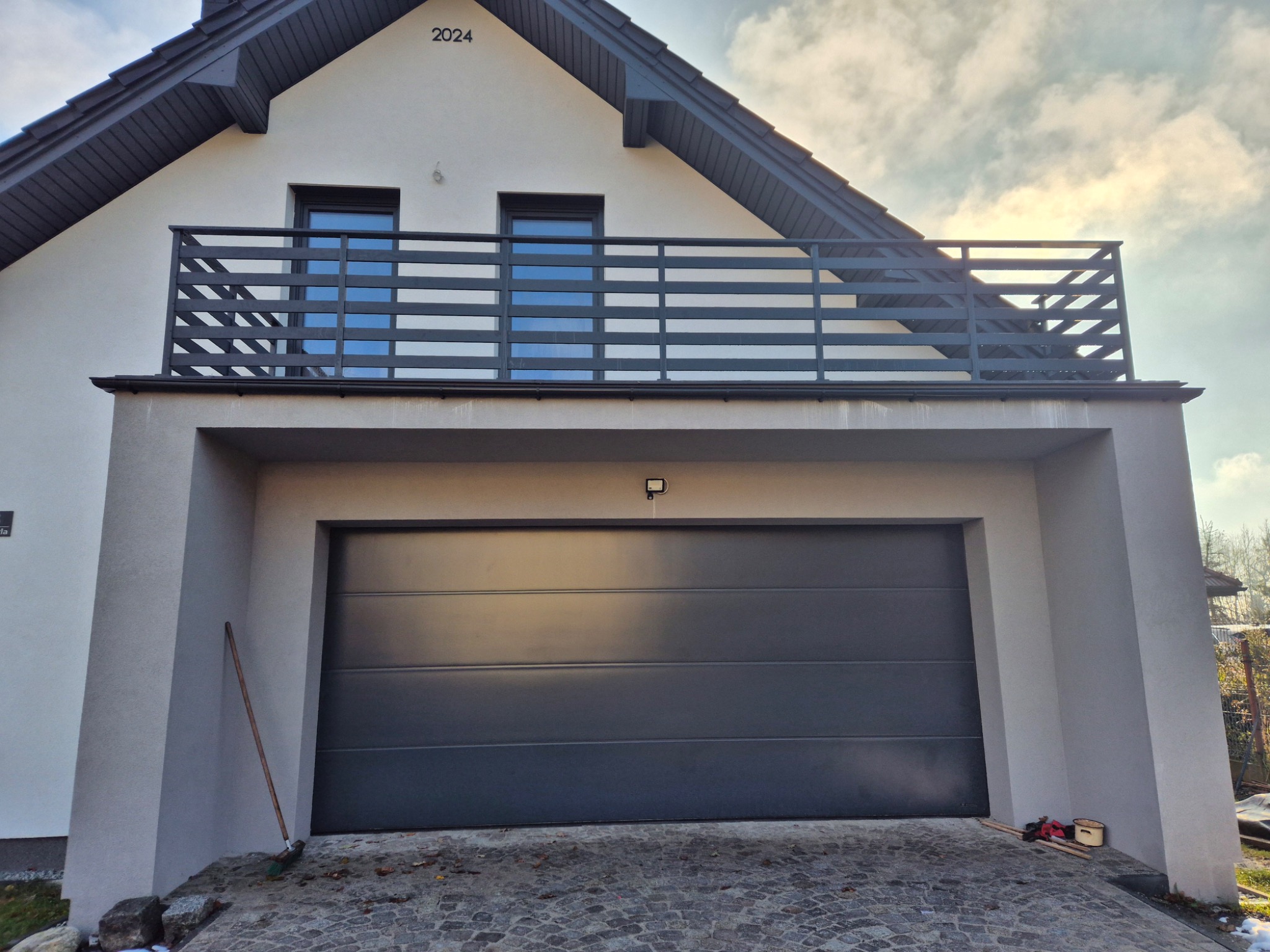 Nowoczesny dom z balkonem zabezpieczonym poziomą, ciemnoszarą konstrukcją, widok na elewację i garaż, kostka brukowa na podjeździe, numer domu '2024' nad balkonem.