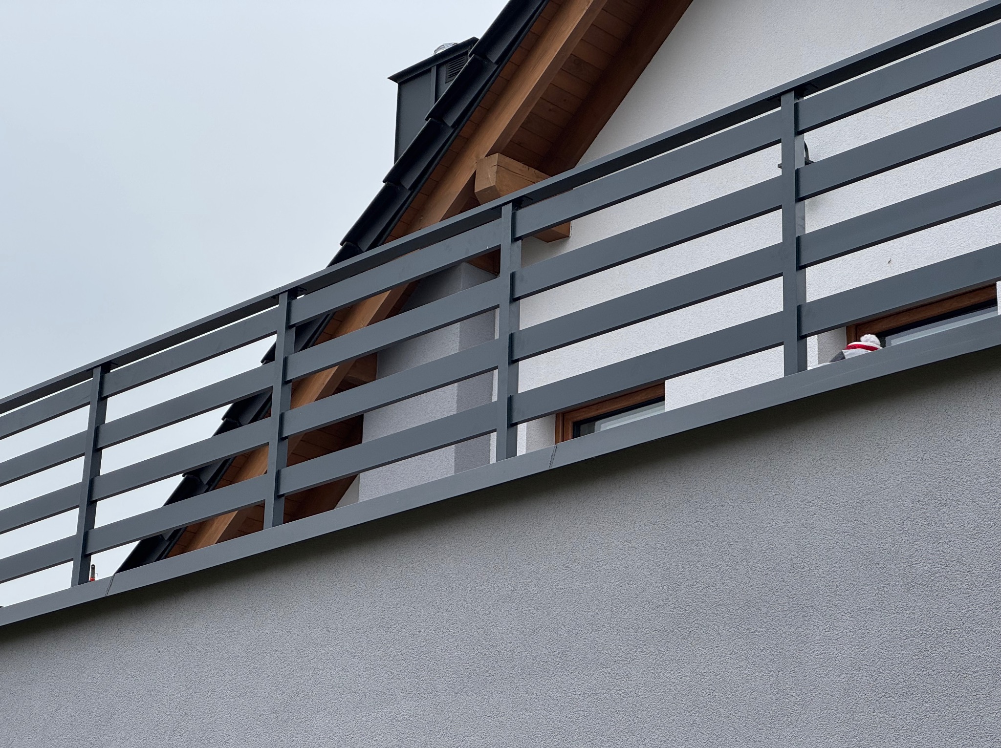 Szara, pozioma balustrada balkonowa z drewnianymi elementami, widok z dołu na tle elewacji i fragmentu dachu domu.
