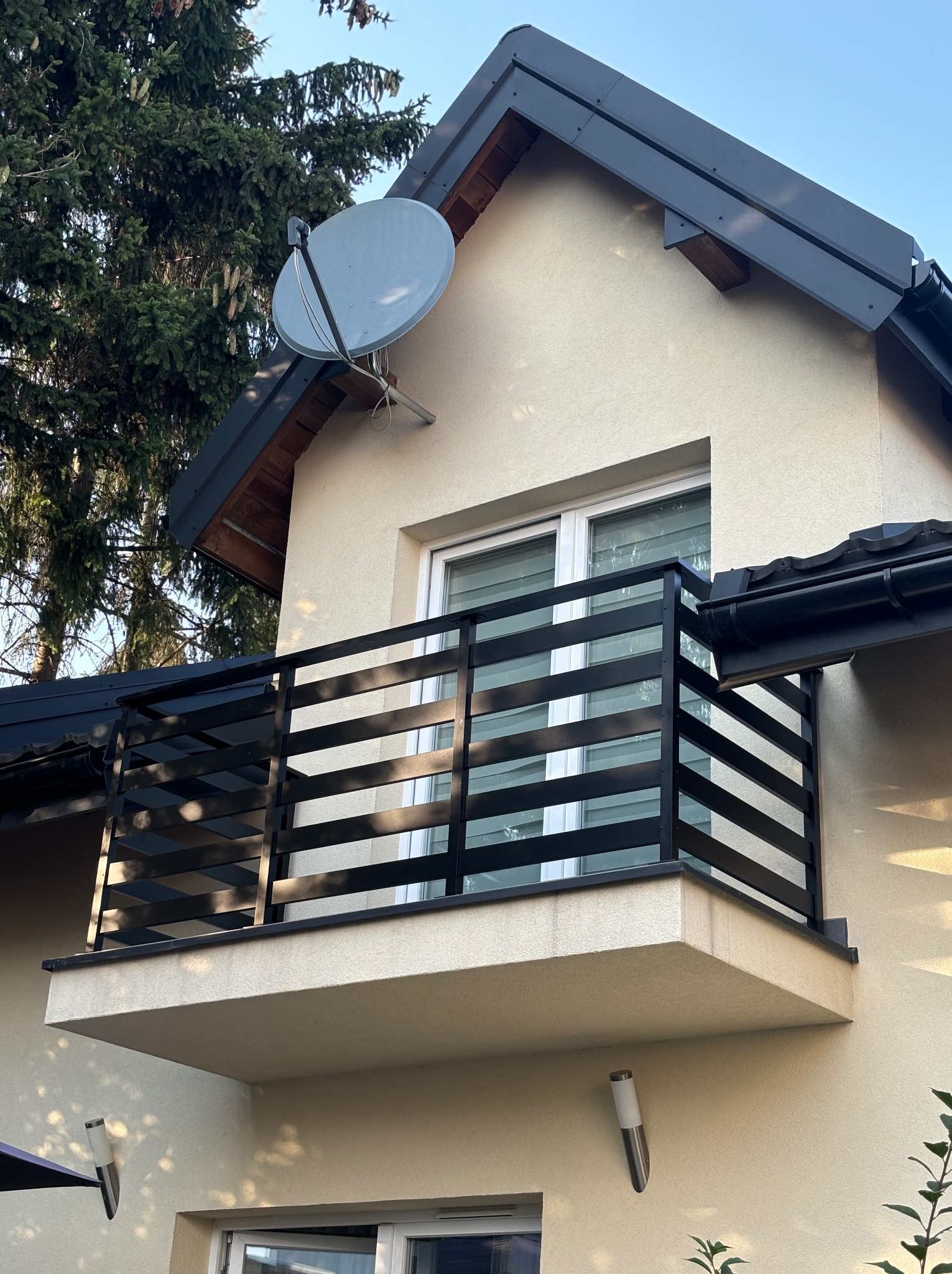 Czarna, pozioma balustrada na balkonie budynku z beżową elewacją i satelitą na dachu, oświetlona popołudniowym słońcem.