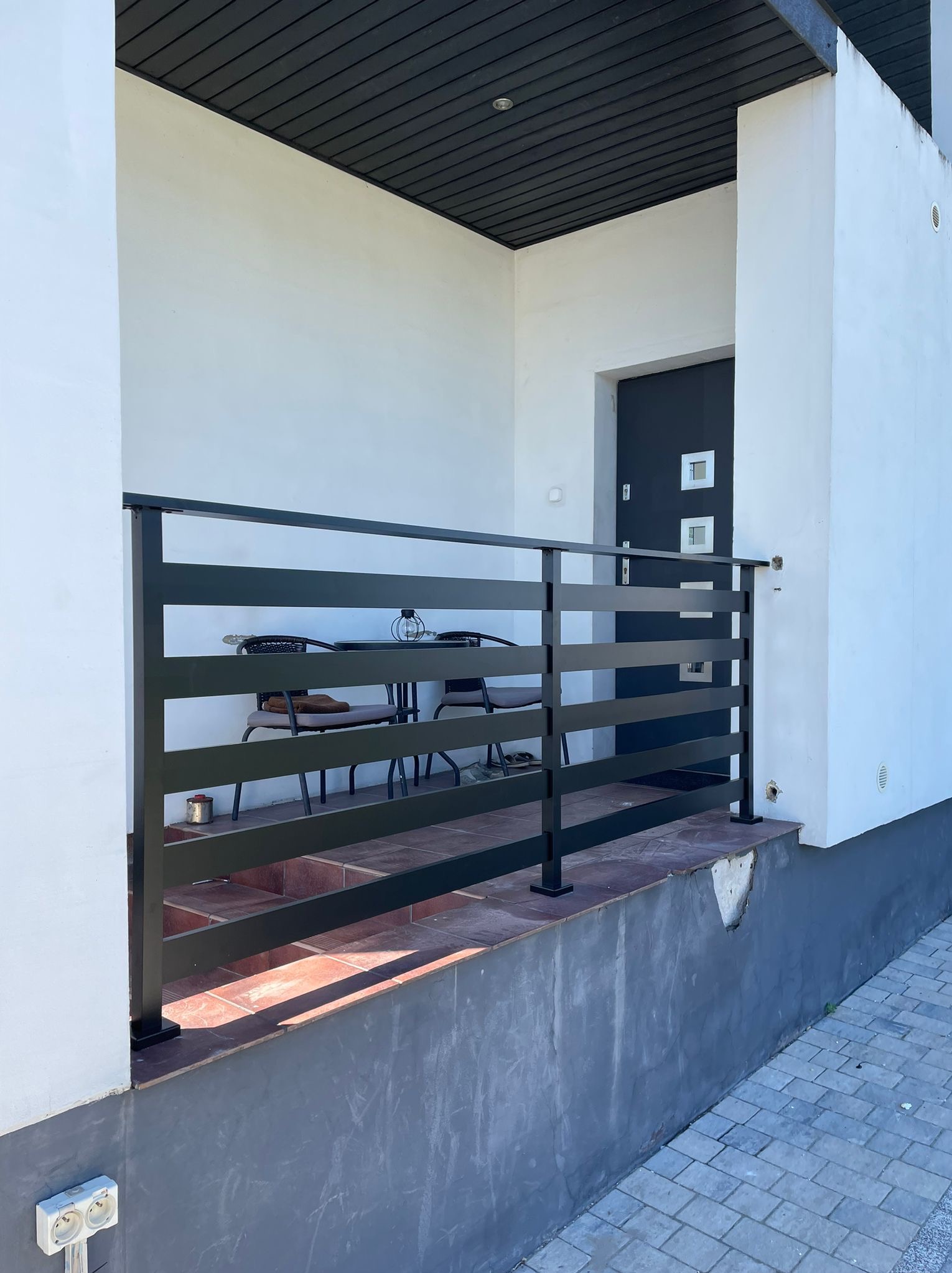 Nowoczesna, czarna balustrada z poziomych profili na tarasie z brązową podłogą, widok na stół i krzesła na zewnątrz budynku z białą elewacją i szarą stolarką.