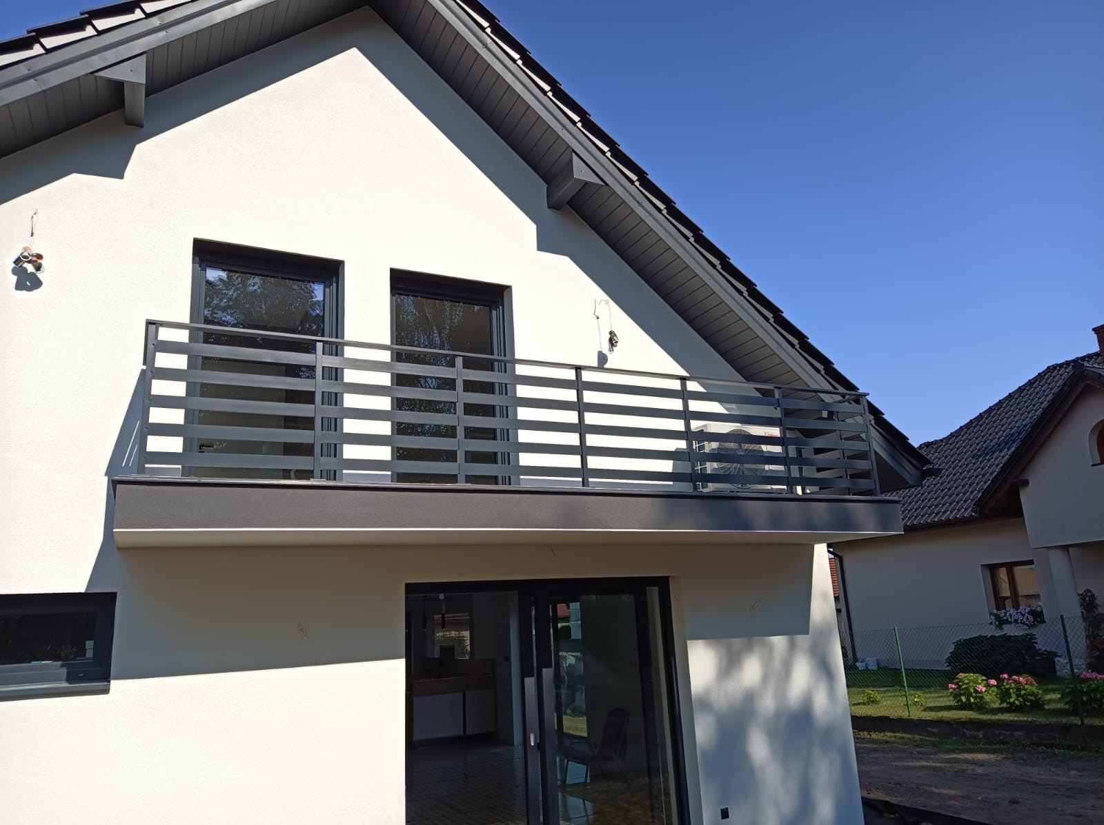 Nowoczesny balkon z szarą, poziomą balustradą na tle białej elewacji domu i błękitnego nieba. Widoczne dwa okna balkonowe i fragment dachu sąsiedniego budynku.