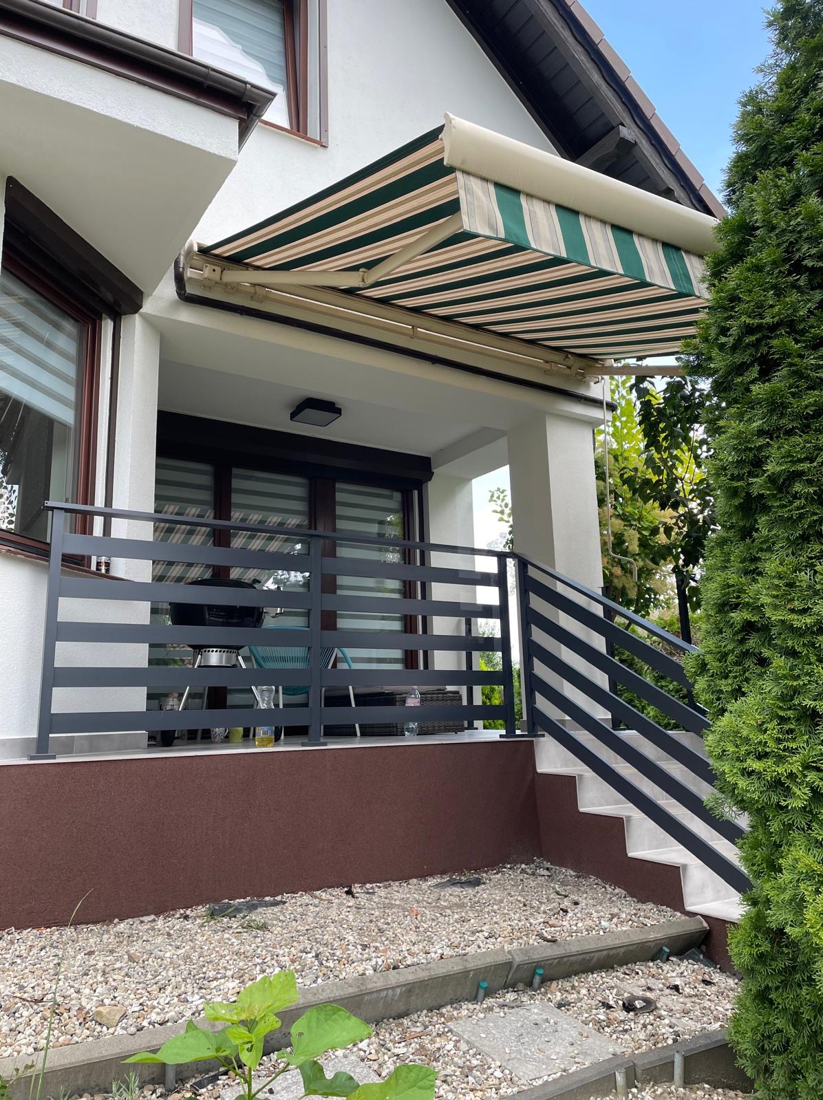 Zadaszony balkon z szarą balustradą poziomą, zewnętrzna roleta w biało-zielone paski, białe filary i schody, brązowa elewacja przyziemia, kamienisty teren.
