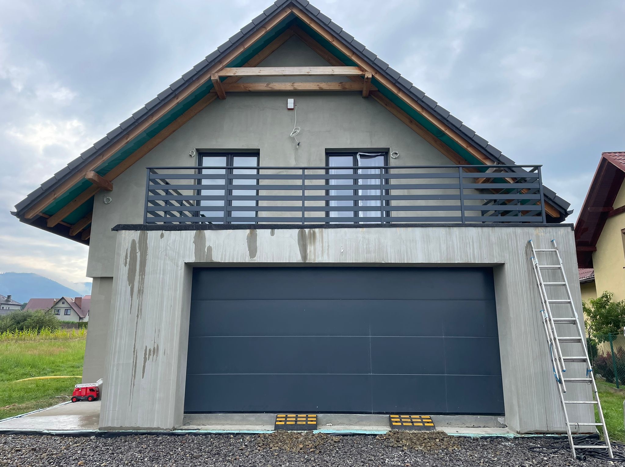 Surowy budynek z balkonem zabezpieczonym metalową barierką, garaż z szarą bramą segmentową, drabina oparta o ścianę, zabawka straż pożarna na podjeździe, widoczne ślady po pracach murarskich...