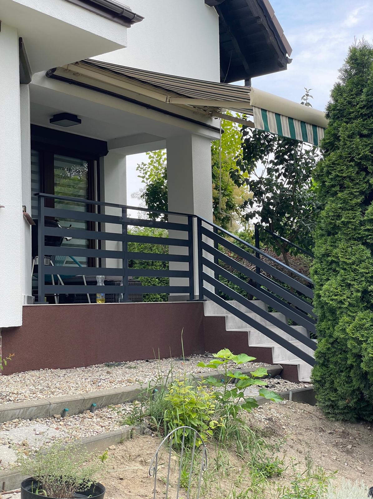 Zewnętrzna, pozioma balustrada w kolorze antracytowym, z prostokątnymi poprzeczkami, zamontowana przy schodach wejściowych do domu z brązową elewacją i markizą w paski.