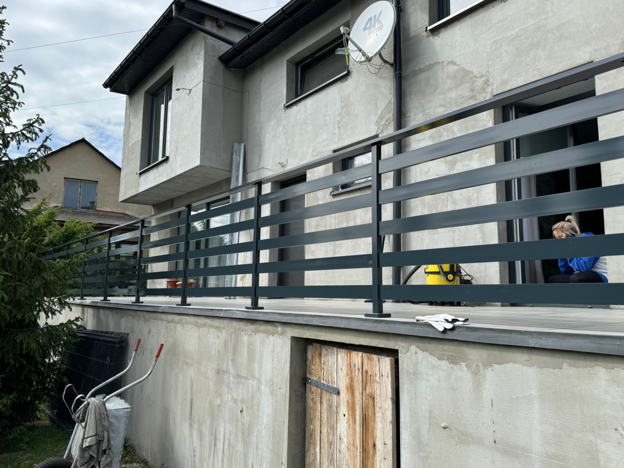Nowoczesna, pozioma, ciemnoszara barierka na tarasie domu z widoczną anteną satelitarną, w tle kobieta siedząca na podłodze tarasu oraz wózek i narzędzia ogrodowe.