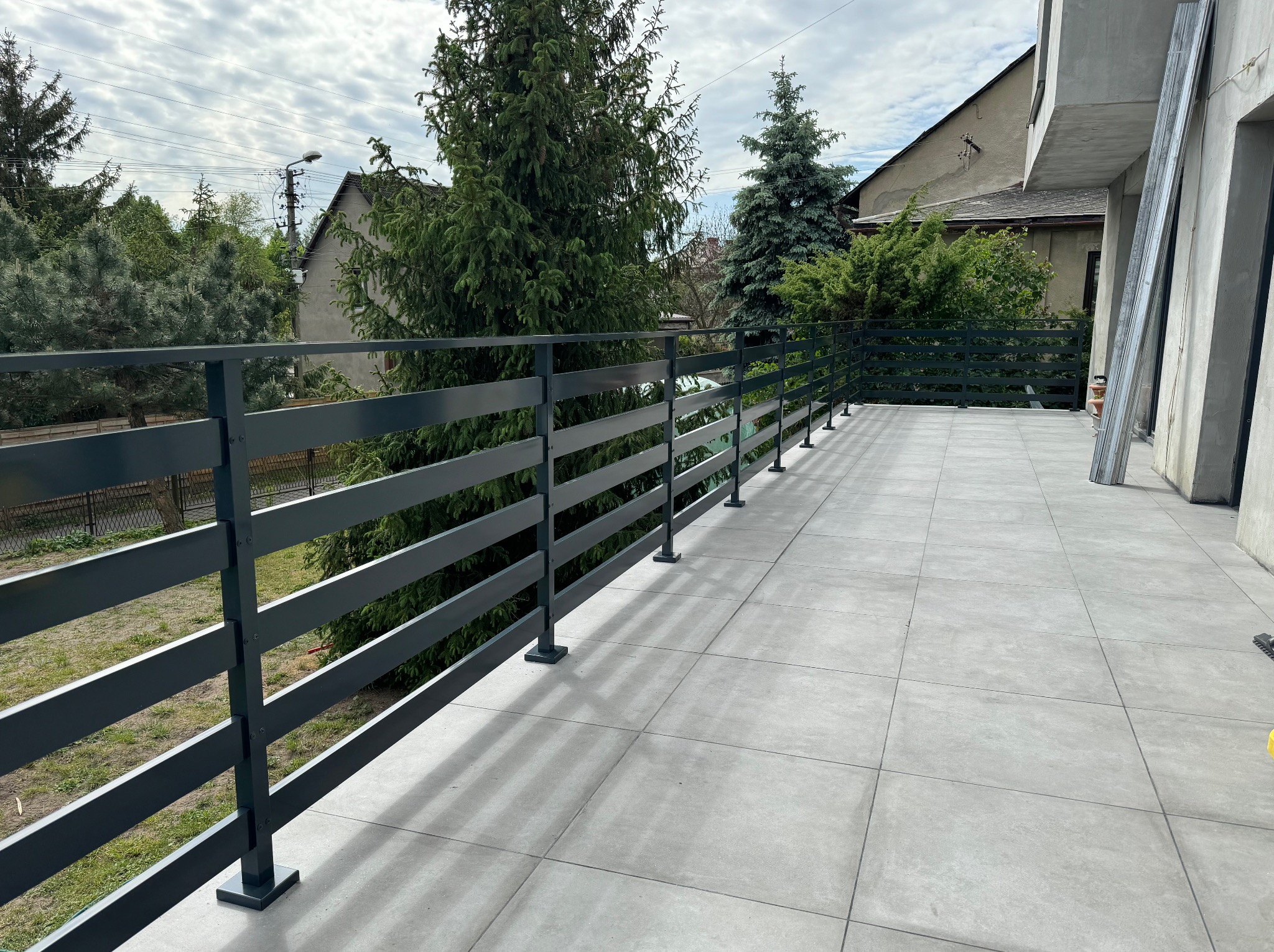 Nowoczesna, pozioma, grafitowa balustrada na tarasie wyłożonym szarą, kwadratową płytką, z widokiem na zieleń ogrodową i zabudowę jednorodzinną w tle.