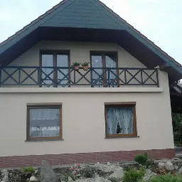 Elewacja domu jednorodzinnego z beżową warstwą wykończeniową, balkon z czarną metalową balustradą w kształcie krzyżyków, czerwona ceglana podmurówka i brązowe ramy okienne.