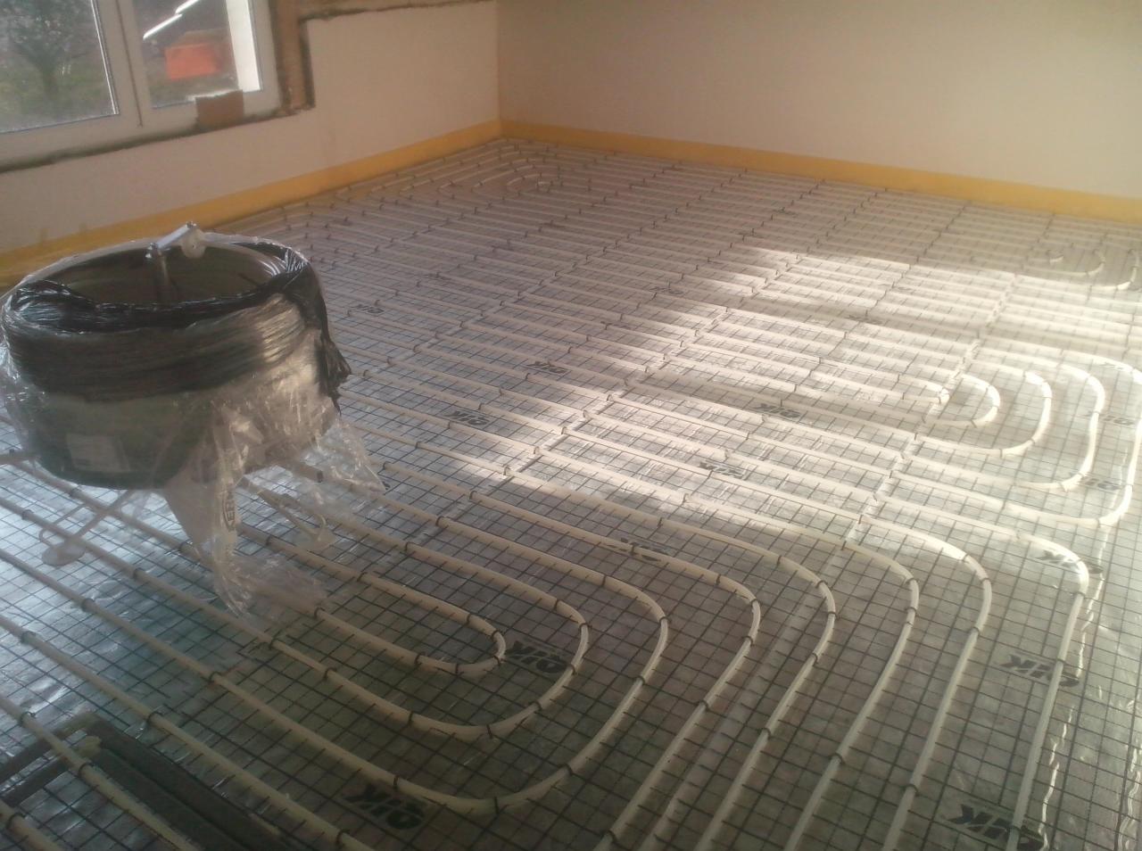 Rozłożona instalacja ogrzewania podłogowego na siatce, z rolką rury w folii w rogu pomieszczenia, widoczne okno i przygotowane ściany do wykończenia.