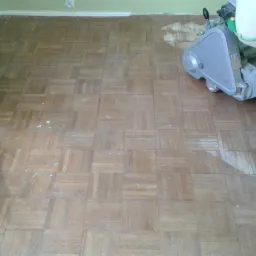 Parkiet w kwadraty częściowo wyszlifowany maszyną, widoczne białe ślady po szpachli na drewnianej podłodze, przy ścianie listwa przypodłogowa.