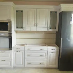 Biała zabudowa kuchenna z piekarnikiem, mikrofalówką i lodówką, widoczne niedokończone blaty bez zlewu i płyty grzewczej, dekoracyjne szklane wstawki w szafkach wiszących.