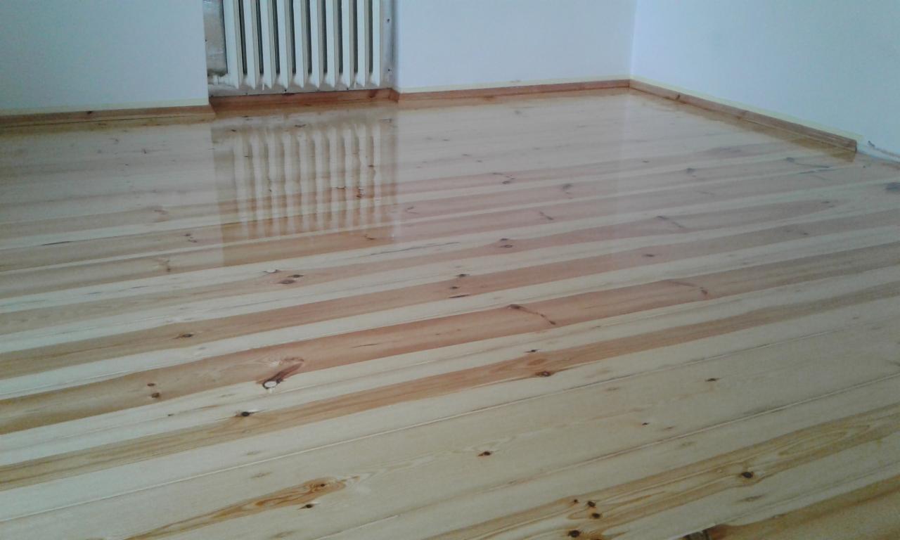 Świeżo położona, lakierowana podłoga z jasnego drewna sosnowego z widocznymi słojami i sękami, odbijająca światło grzejnika w rogu pokoju.