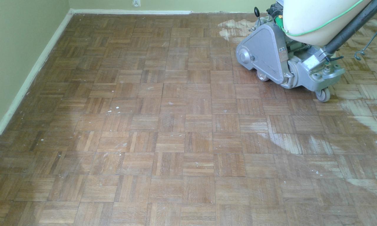 Parkiet w kwadraty częściowo wyszlifowany maszyną, widoczne białe ślady po szpachli na drewnianej podłodze, przy ścianie listwa przypodłogowa.
