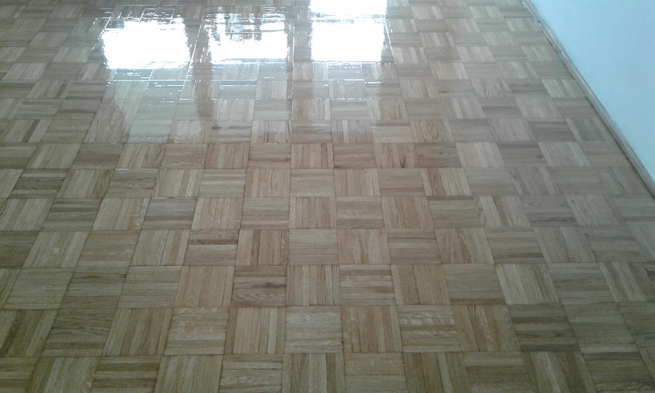Świeżo polakierowana, błyszcząca podłoga parkietowa ułożona w kwadraty, z widocznym odbiciem okna.