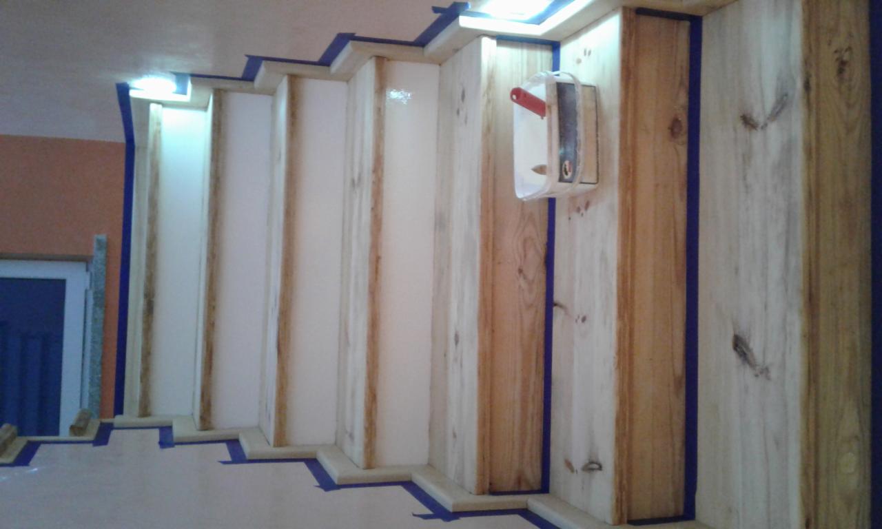 Fragment drewnianych schodów w trakcie malowania, zabezpieczonych niebieską taśmą malarską, widoczny pojemnik z narzędziami na ścianie.