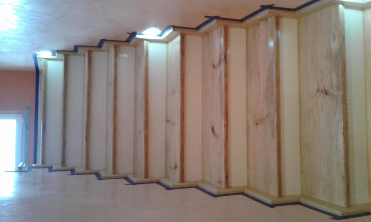 Wnętrze w trakcie montażu drewnianej konstrukcji, widoczne pionowe elementy z jasnego drewna z ciemnymi sękami oraz niebieska taśma malarska zabezpieczająca krawędzie, sufit z widocznymi lampami.