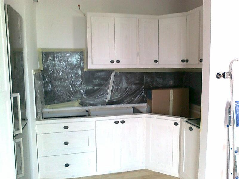 Biała, drewniana zabudowa kuchenna z czarnymi uchwytami, zabezpieczona folią malarską podczas prac wykończeniowych, widoczny karton w tle.