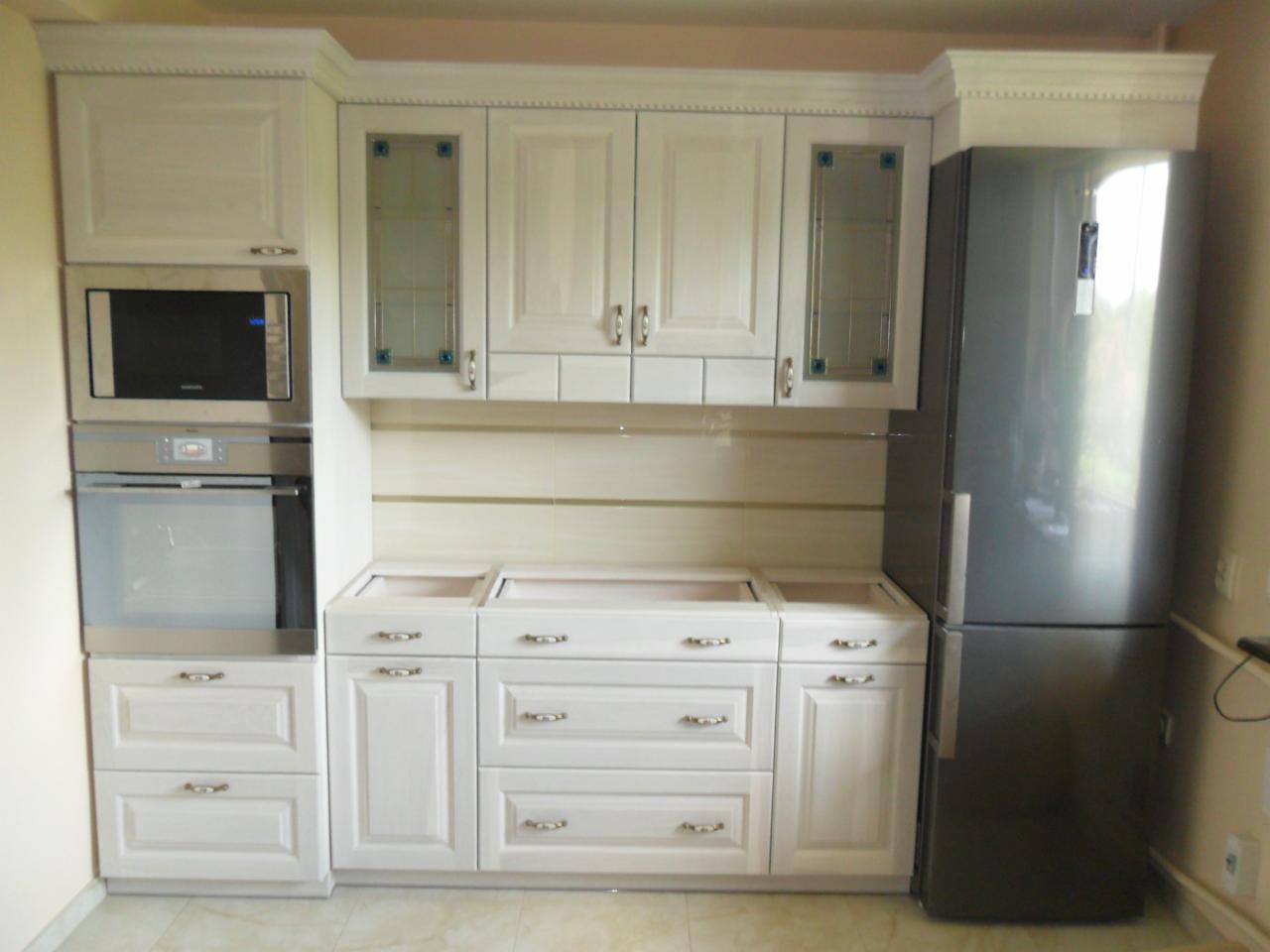 Biała zabudowa kuchenna z piekarnikiem, mikrofalówką i lodówką, widoczne niedokończone blaty bez zlewu i płyty grzewczej, dekoracyjne szklane wstawki w szafkach wiszących.