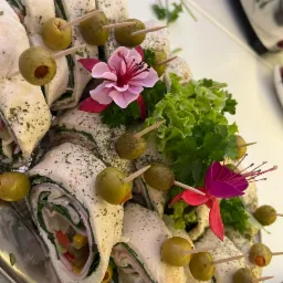 Elegancko ułożone roladki z tortilli z szynką, warzywami i oliwkami na wykałaczkach, udekorowane sałatą i kwiatami na srebrnej tacy.