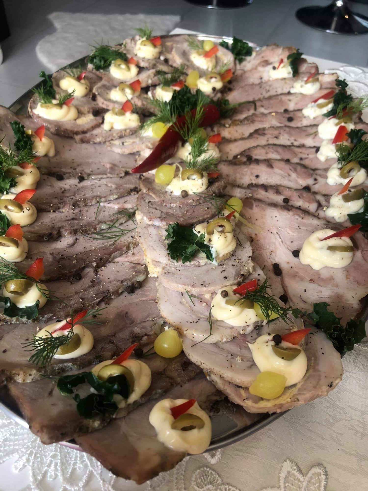 Plater z plastrami pieczonego mięsa, udekorowany kremem, oliwkami, papryką i koperkiem, ułożony na białym obrusie.