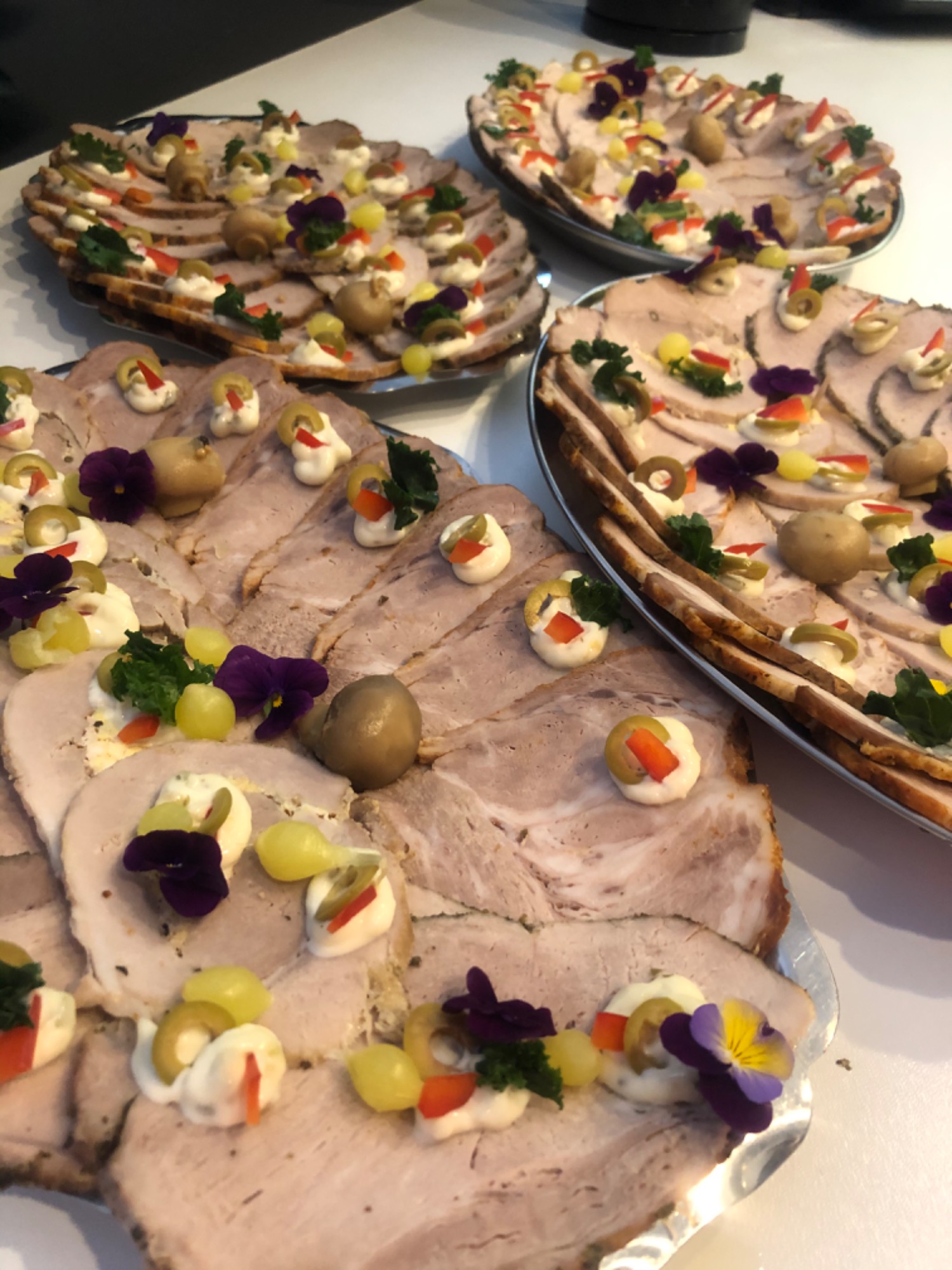 Trzy owalne półmiski z plastrami pieczonego mięsa, dekorowane kremowym sosem, oliwkami, papryką, winogronami, grzybami i fioletowymi bratkami.