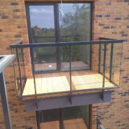Nowoczesny balkon z drewnianą podłogą i szklaną balustradą, zamontowany na elewacji z cegły klinkierowej. W szybie okna odbija się osoba robiąca zdjęcie.