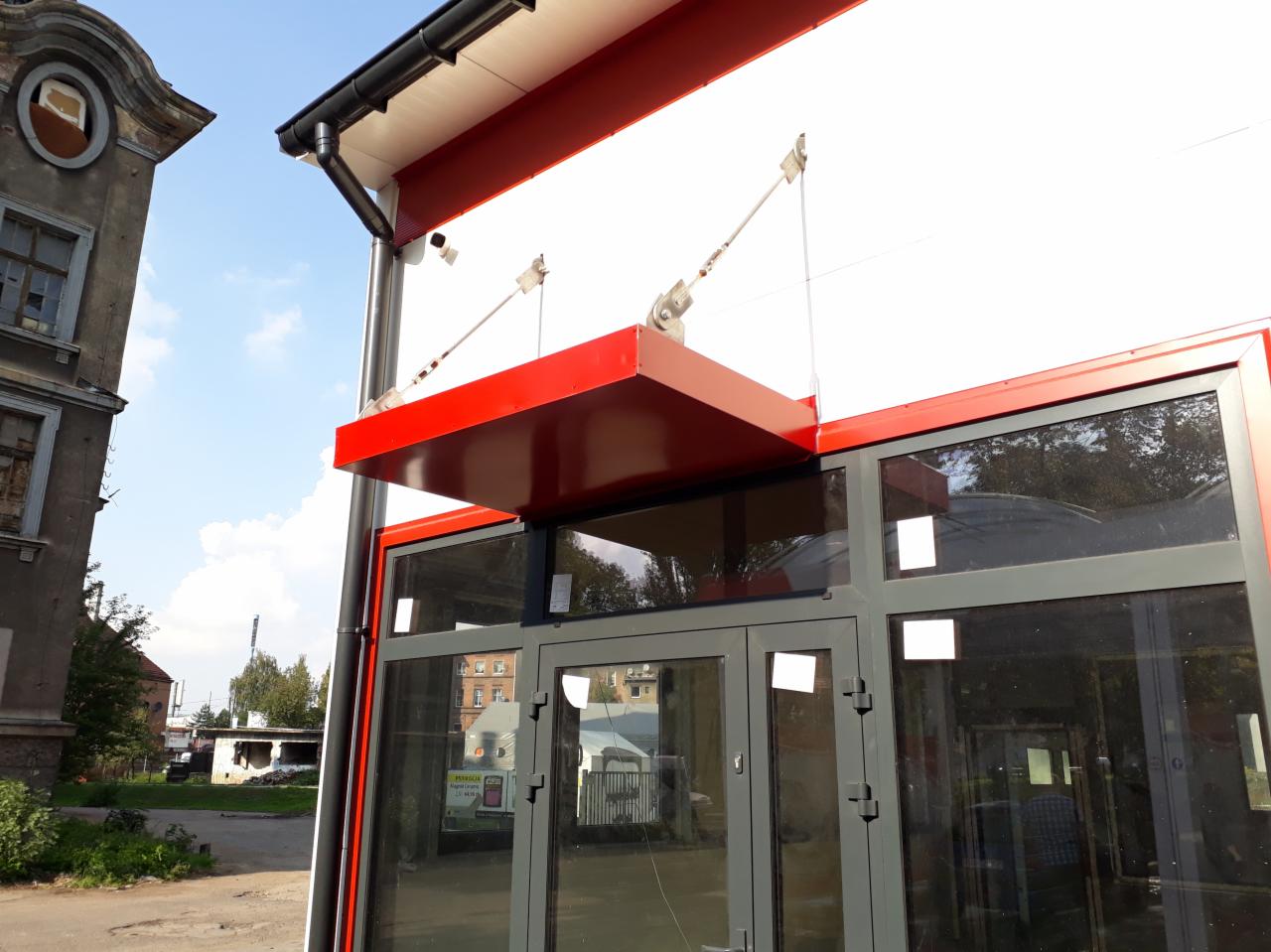 Nowoczesne wejście do budynku z przeszklonymi drzwiami i czerwonym zadaszeniem wspartym na metalowych linkach, widoczne fragmenty elewacji i sąsiedniego, starszego budynku.