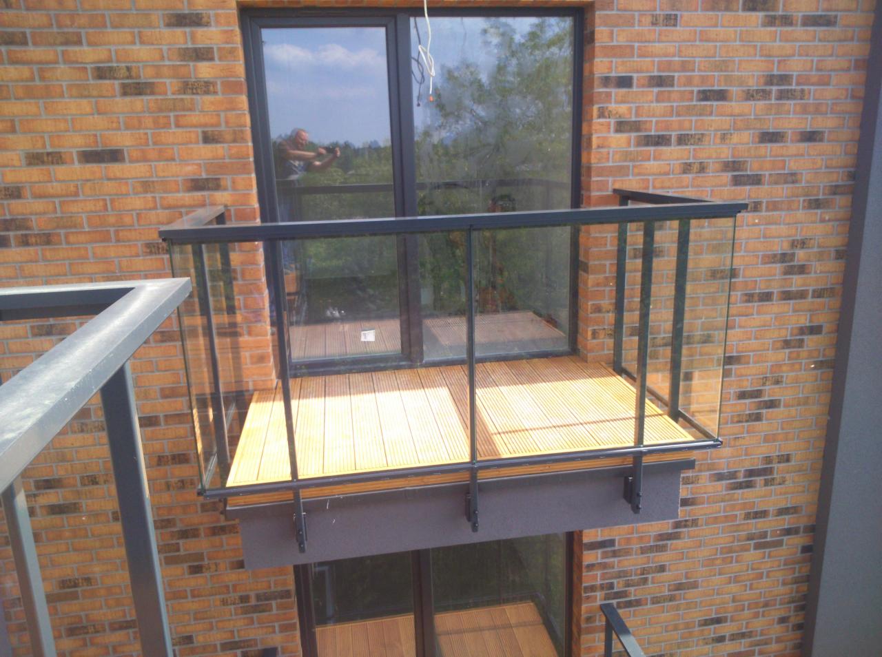 Nowoczesny balkon z drewnianą podłogą i szklaną balustradą, zamontowany na elewacji z cegły klinkierowej. W szybie okna odbija się osoba robiąca zdjęcie.